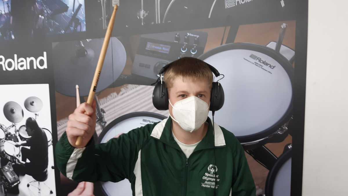 Alex wearing headphones and a mask and holding a drum stick. Behind him in a poster with a drum kit on it.