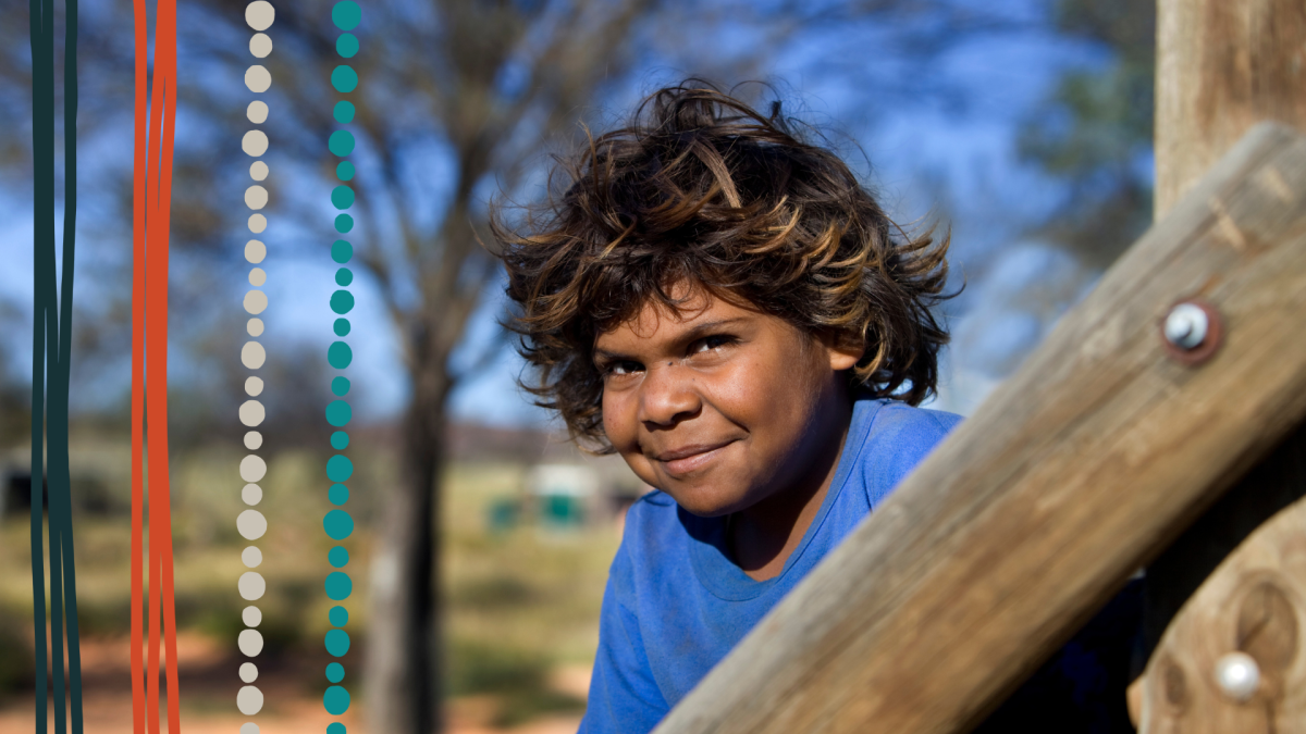 A young Aboriginal boy with curly brown hair wearing a blue t-shirt smiles at the camera.