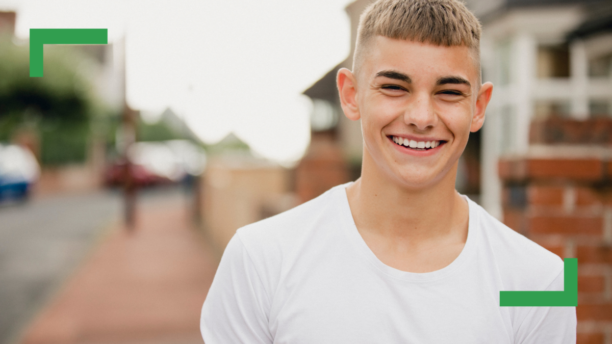 A teenage boy with blonde hair, wearing a white T-shirt, stands outside, smiling at the camera.