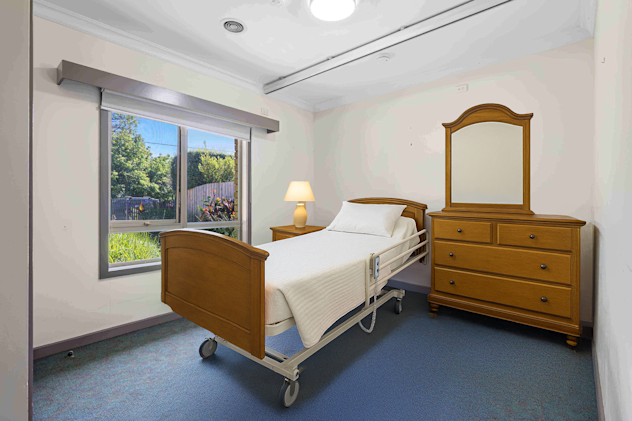 Neat and simple bedroom featuring a hospital-style single bed with a wooden headboard and footboard, a bedside table with a lit lamp, and a wooden dresser with an attached mirror. The room has a large window with a garden view and blue carpet flooring.