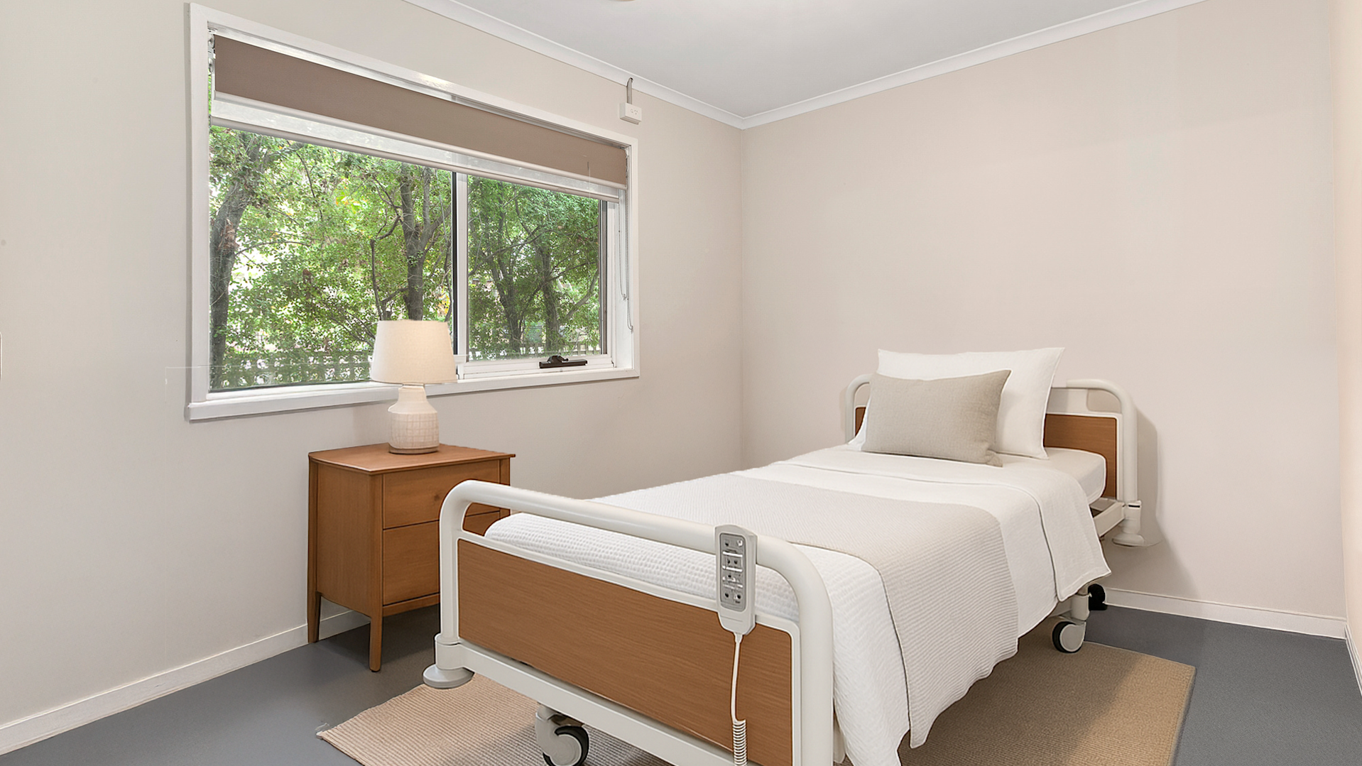 Simple and clean bedroom with a single adjustable hospital-style bed, light beige walls, a wooden bedside table with a lamp, and a large window overlooking green trees.
