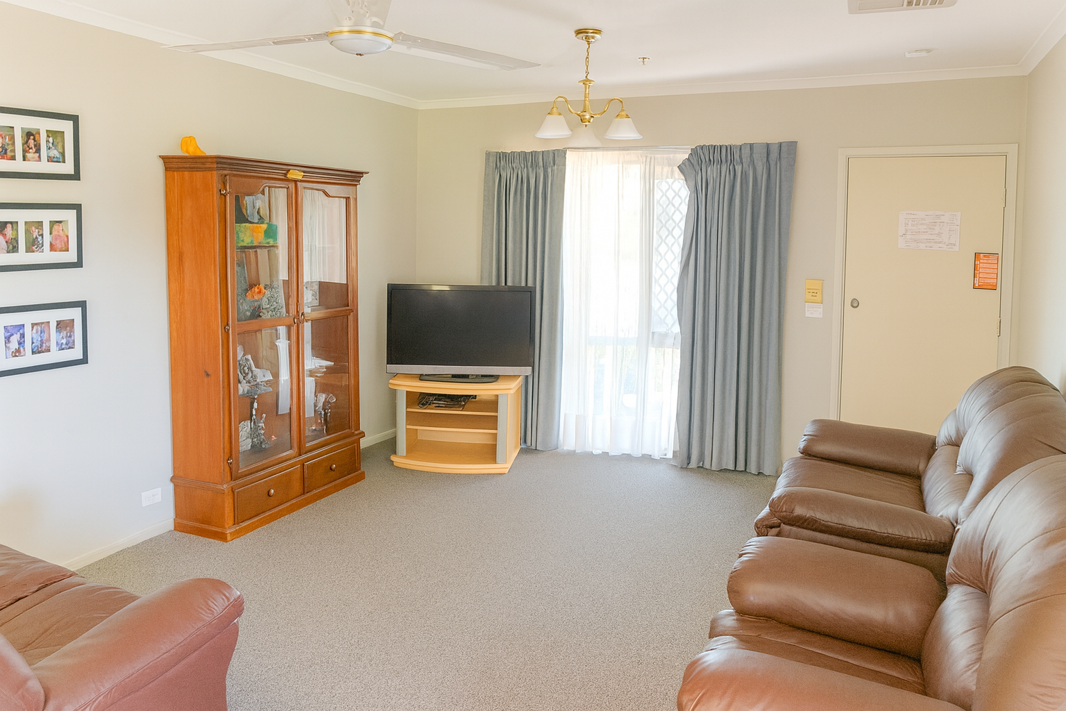 A bright and welcoming living room with light walls, brown leather sofas, a glass display cabinet, and a TV on a wooden stand. Natural light filters through large curtains, creating a warm and inviting atmosphere.