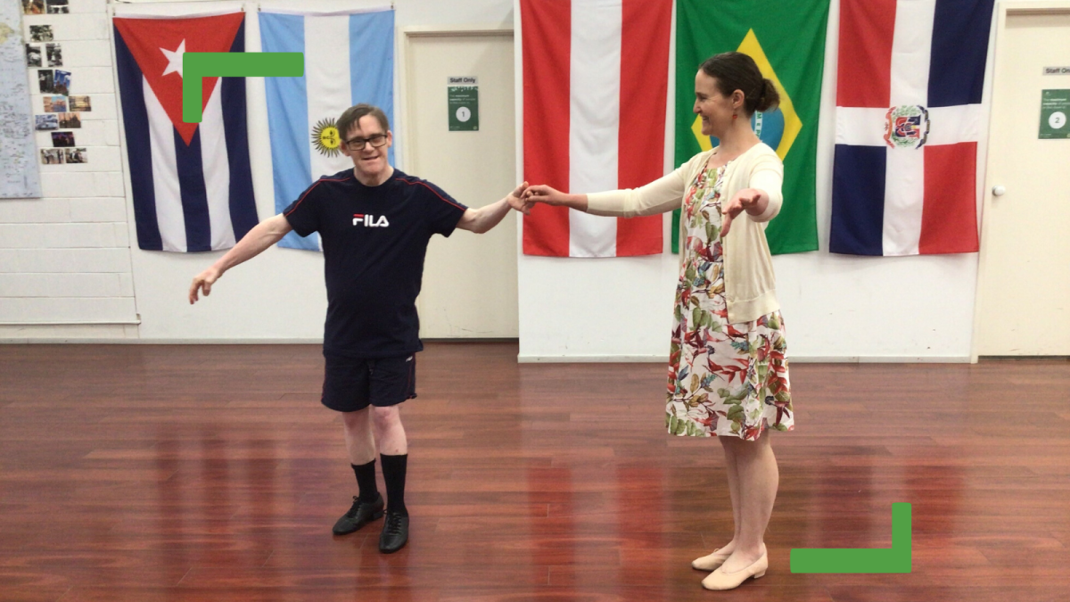 Damien taking dancing lessons. He is wearing a black tshirt, black shorts and black shoes. He is holding the hand of the dance teacher. There are flags in the background.