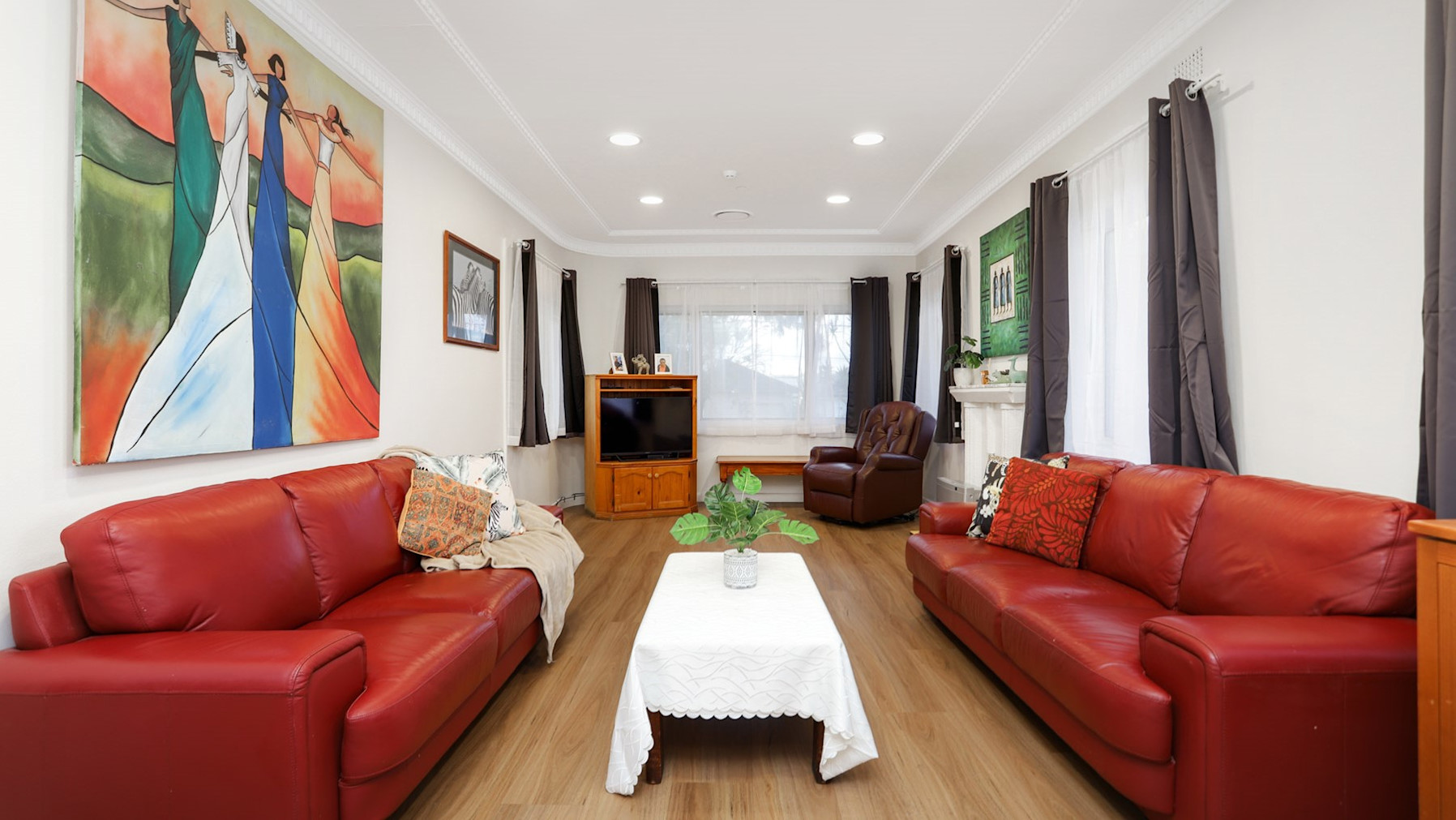 Loungeroom with timber flooring two, large three seater red couches, an armchair, coffee table, television, colourful artwork and curtains.