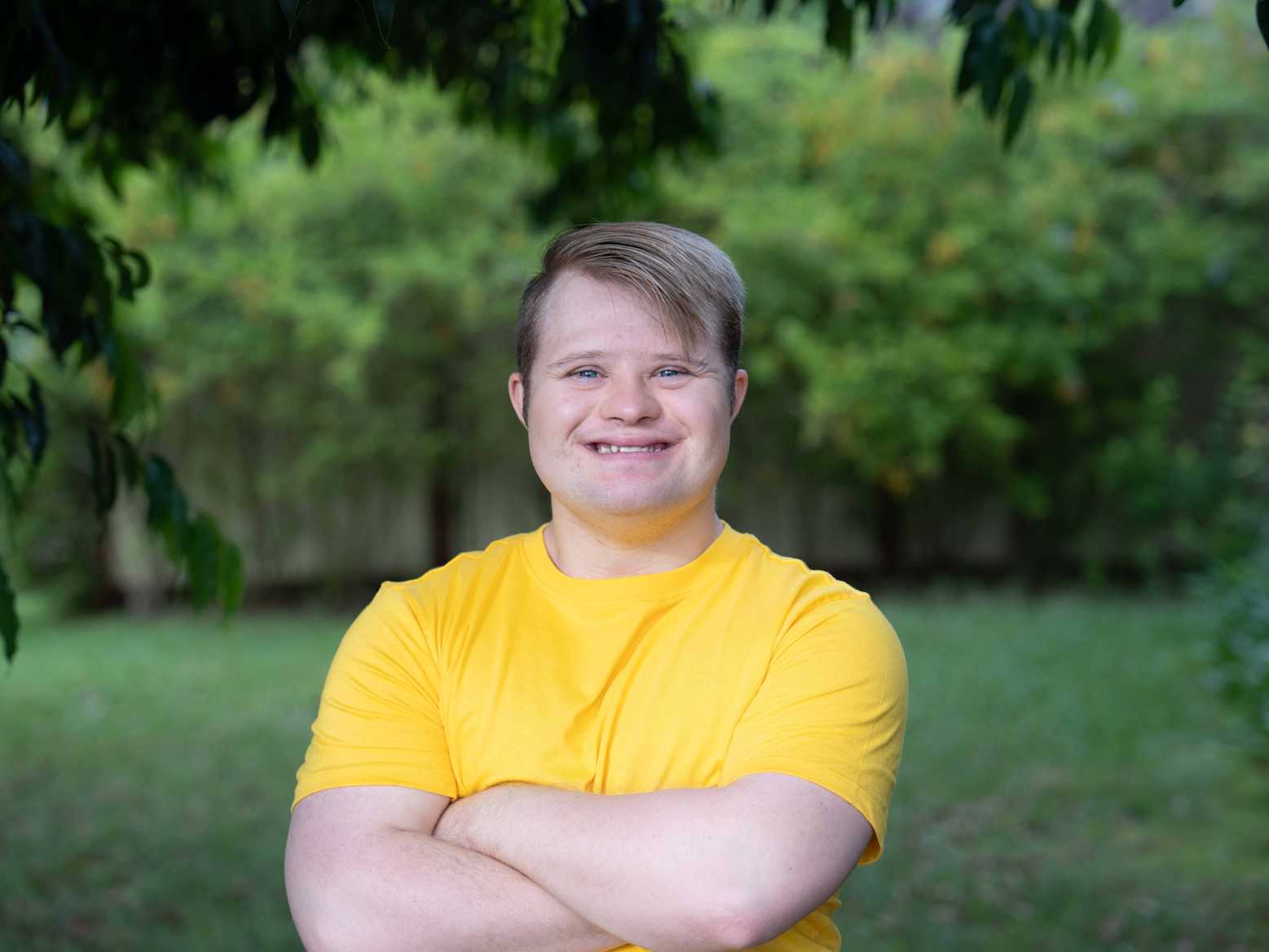 Lee is wearing a bright yellow t-shirt and standing with his arms crossed. There are trees and grass in the background.