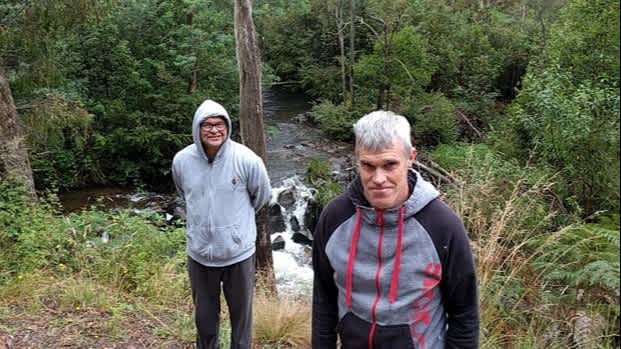 Two men wearing hoodies stand outdoors in a forest smiling at the camera.