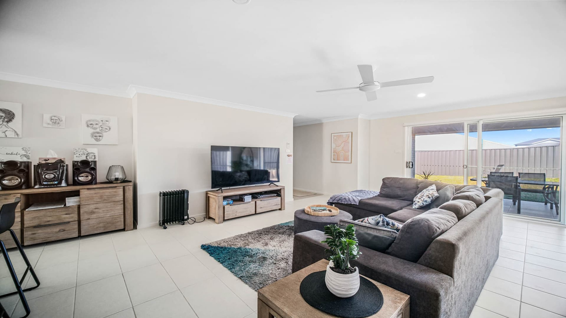 Loungeroom has white tiled floor, brown couches in an L shape, blue, grey and white rug, wooden wall units with sound equipment, facing a television.