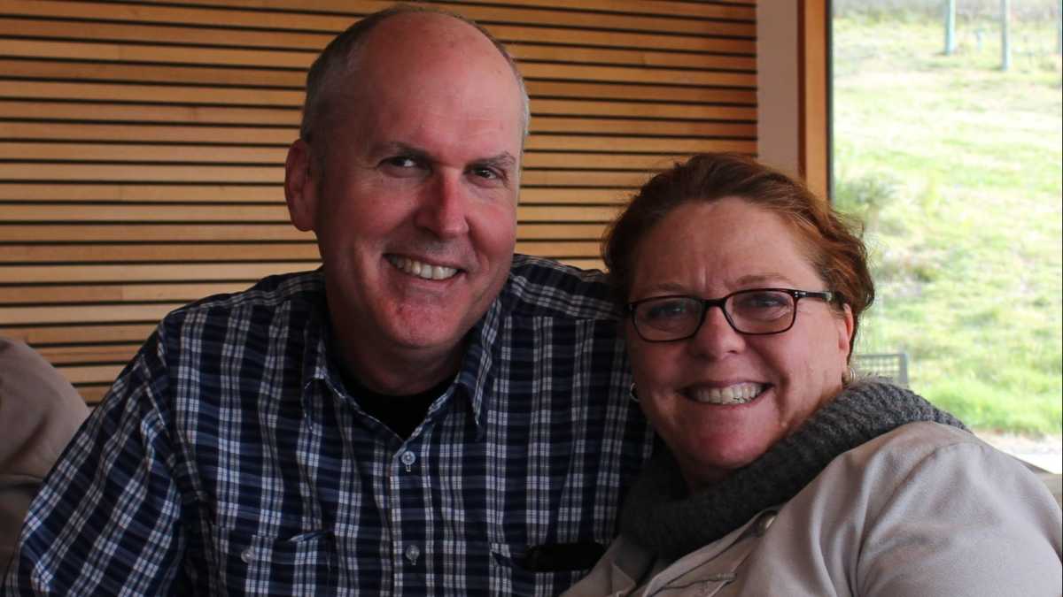 Sandra and Trev smiling into the camera. Sandra is wearing glasses and Trev has a checked shirt on.