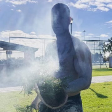 Kurt Fuller, a Wiradjuri man and Life Without Barriers Case Manager wearing white body and face paint holds a bundle of foliage that is smoking.