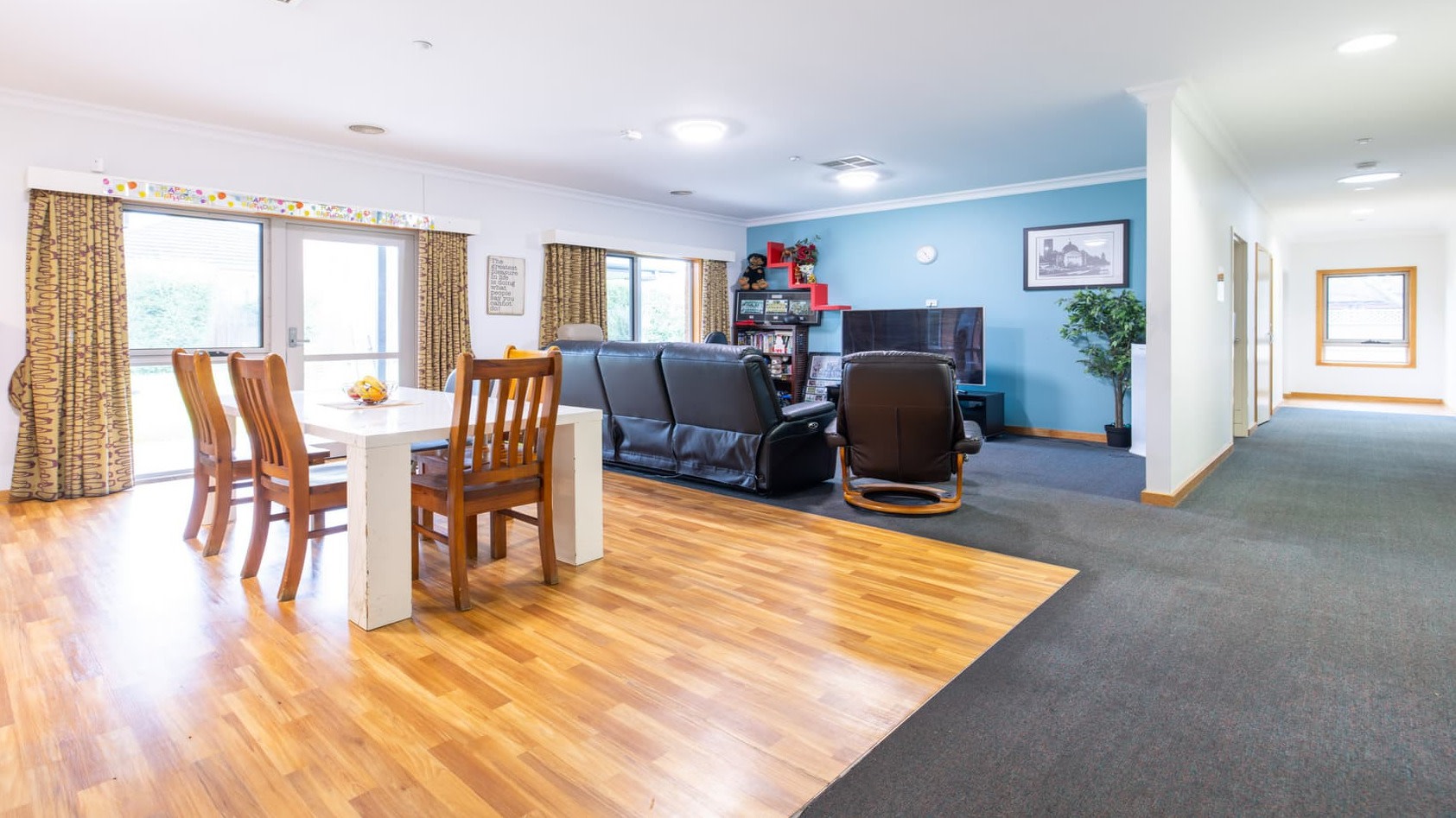 Open-plan living and dining area in a residential home featuring wooden flooring in the dining space with a white table and wooden chairs, transitioning to carpet in the lounge area with black leather sofas, a large TV, bookshelves, and a blue accent wall; a hallway with doors and a window extends to the right.