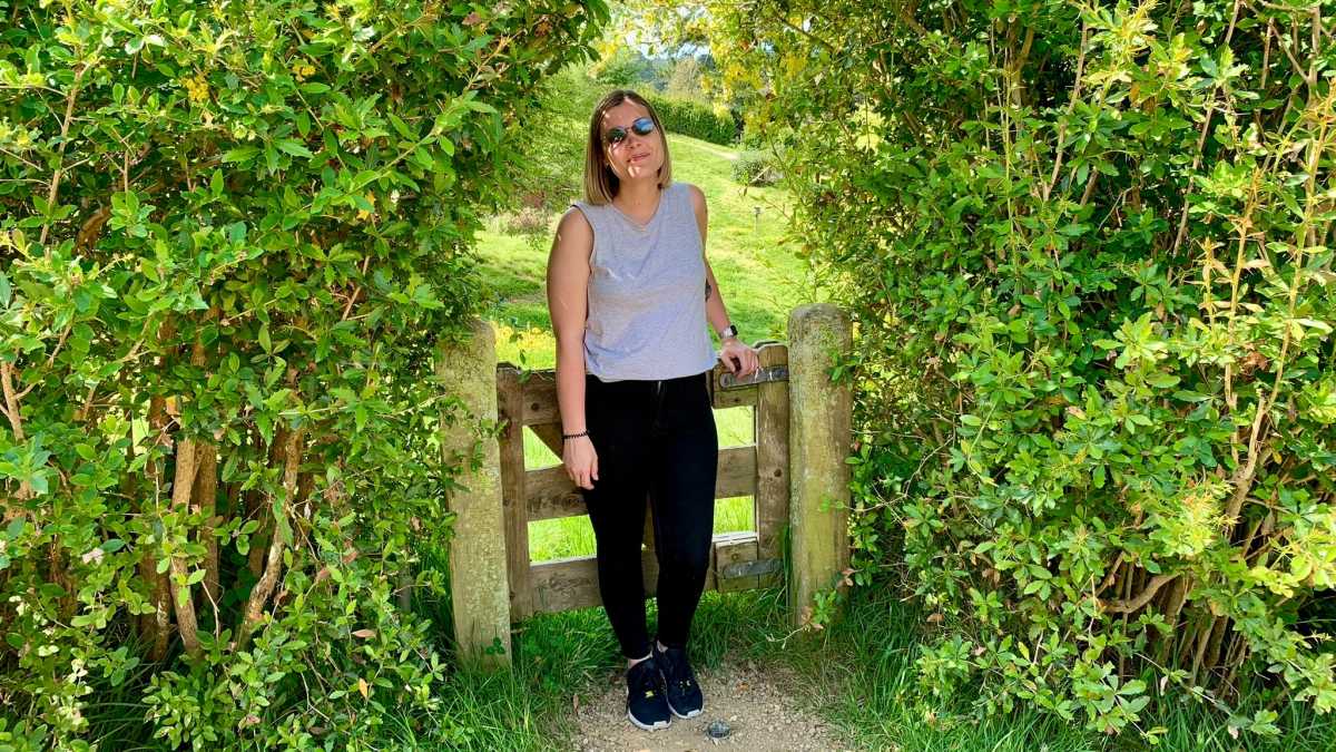 Ashleigh standing outside in front of a gate between two large bushes wearing black pants and a grey singlet top.