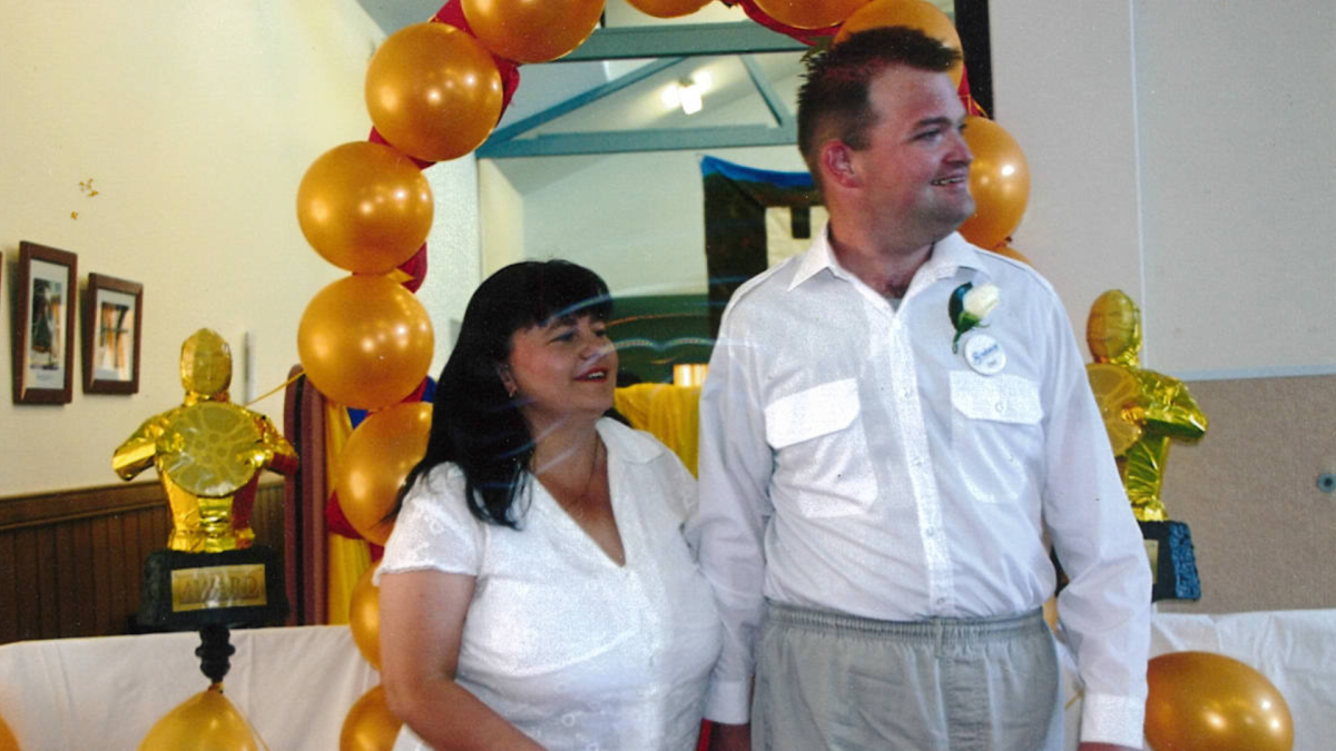 Daniel dressed in a white shirt and slacks standing with a woman with shoulder length brown hair in a white shirt in front of a balloon arch.