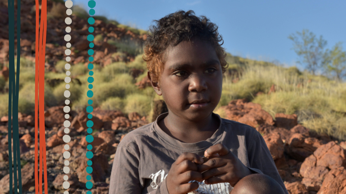 A young Aboriginal boy is sitting outdoors on red rock and grass looking off to the side. He has short curly brown hair and his wearing a grey long sleeved shirt.