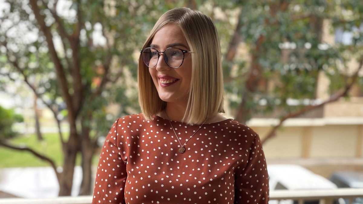 Emily Reaper has short blonde hair and is wearing a brown dress with white spots and glasses.