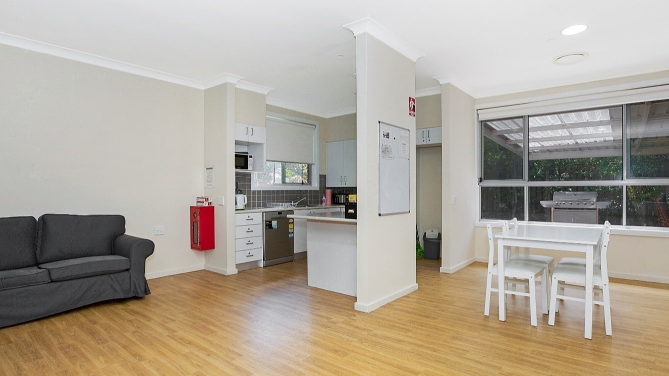 View of open living and dining area with a kitchen in the corner. Floor boards and white walls, dark grey lounge.