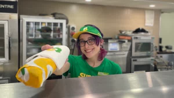 Tiara wearing glasses and a green subway shirt and hat smiling at the camera and holding out a wrapped up sandwich