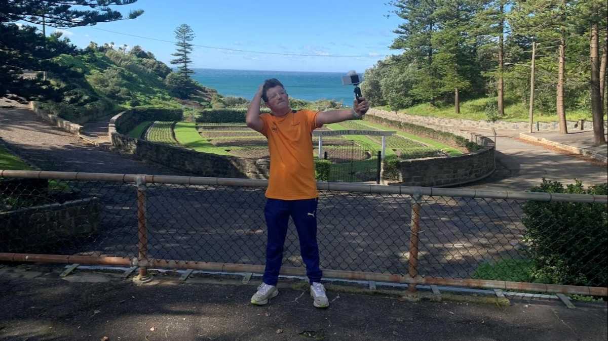 Patrick is wearing blue pants and an orange t-shirt. He stands outdoors in a park holding a camera and taking a photo of himself.