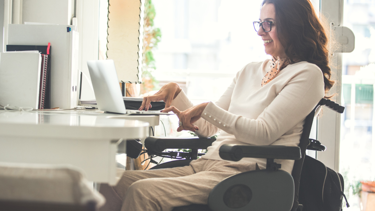 A woman with long brown hair wearing glasses and a cream shirt and pants is sitting in a wheelchair at a desk using a laptop.