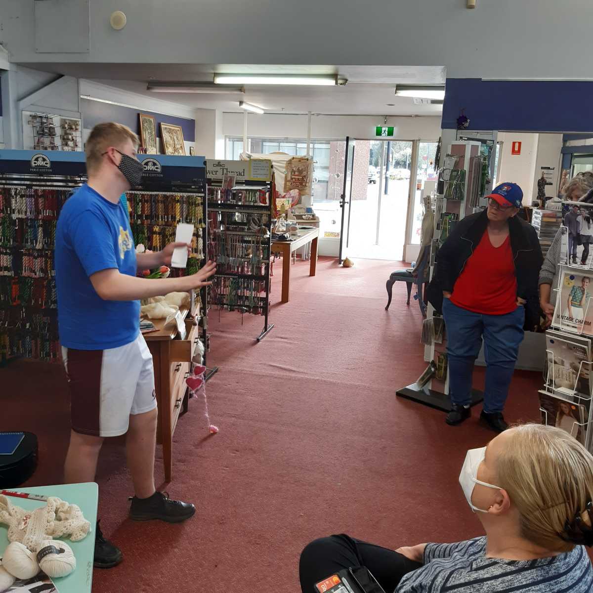 Alex wearing a mask, blue t-shirt and white shorts performing a magic trick to an audience in Hand to Hand Crafts.