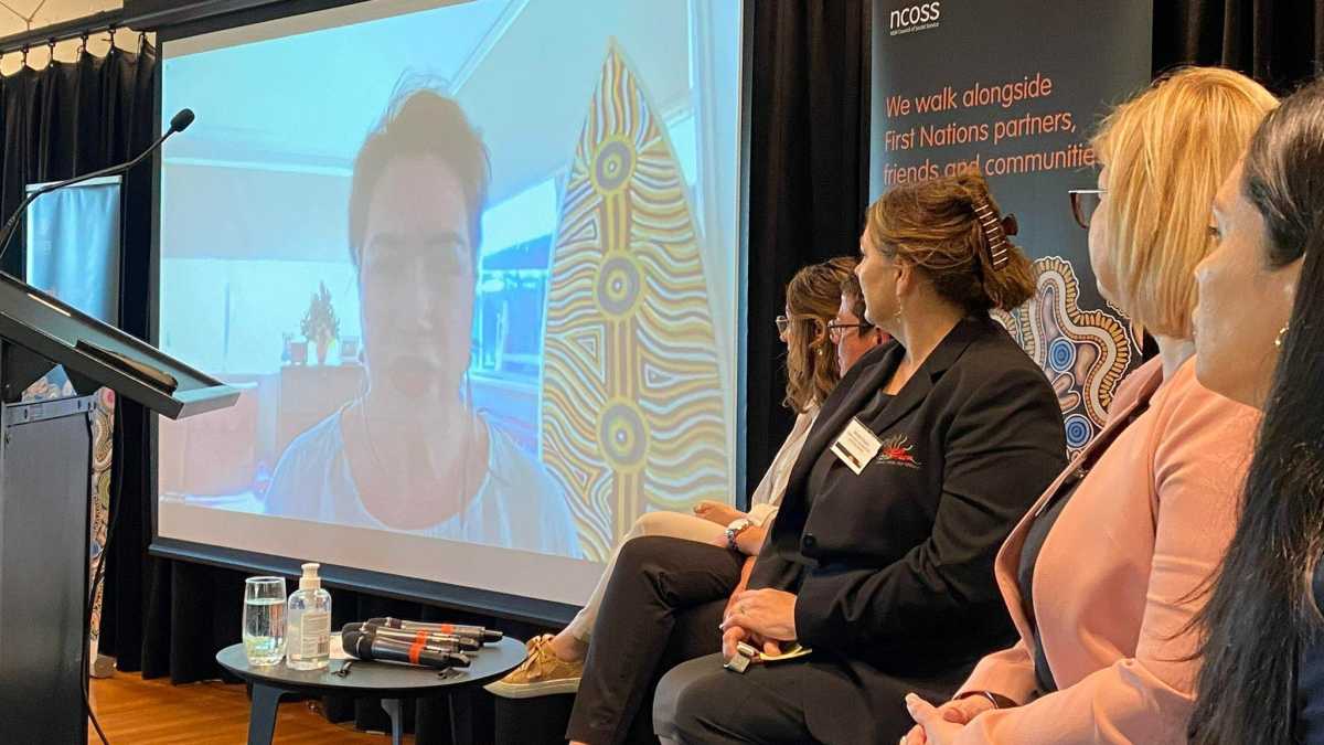 Four women sitting on stage facing a screen where CEO SNAICC Catherine Liddle is speaking.