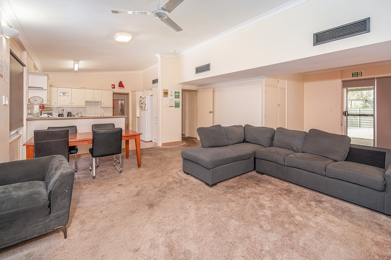 View of a large open-space carpeted lounge room with large grey-seater lounge with four seater dining table and kitchen in the background.