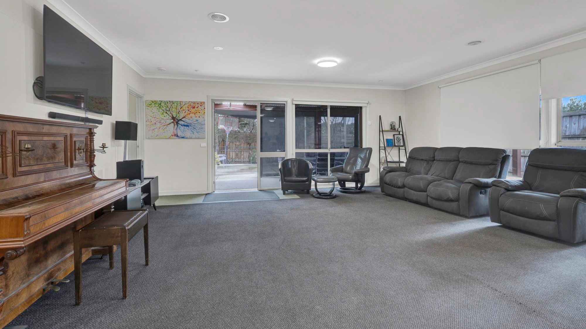 Comfortable living room with a charcoal leather sofa, armchairs, and piano, opening to the outdoor area through glass sliding doors.
