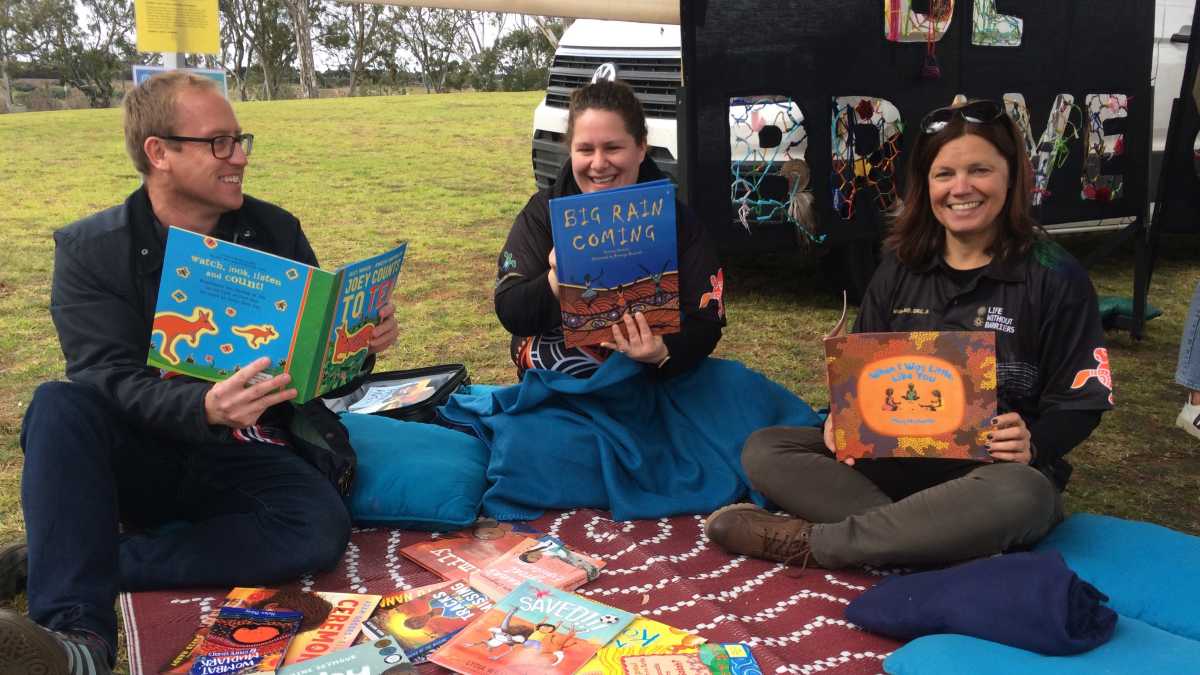 A man and two women sit on a rug with a pile of picture books. They are holding picture books and sit in front of a sign that says Be Brave.