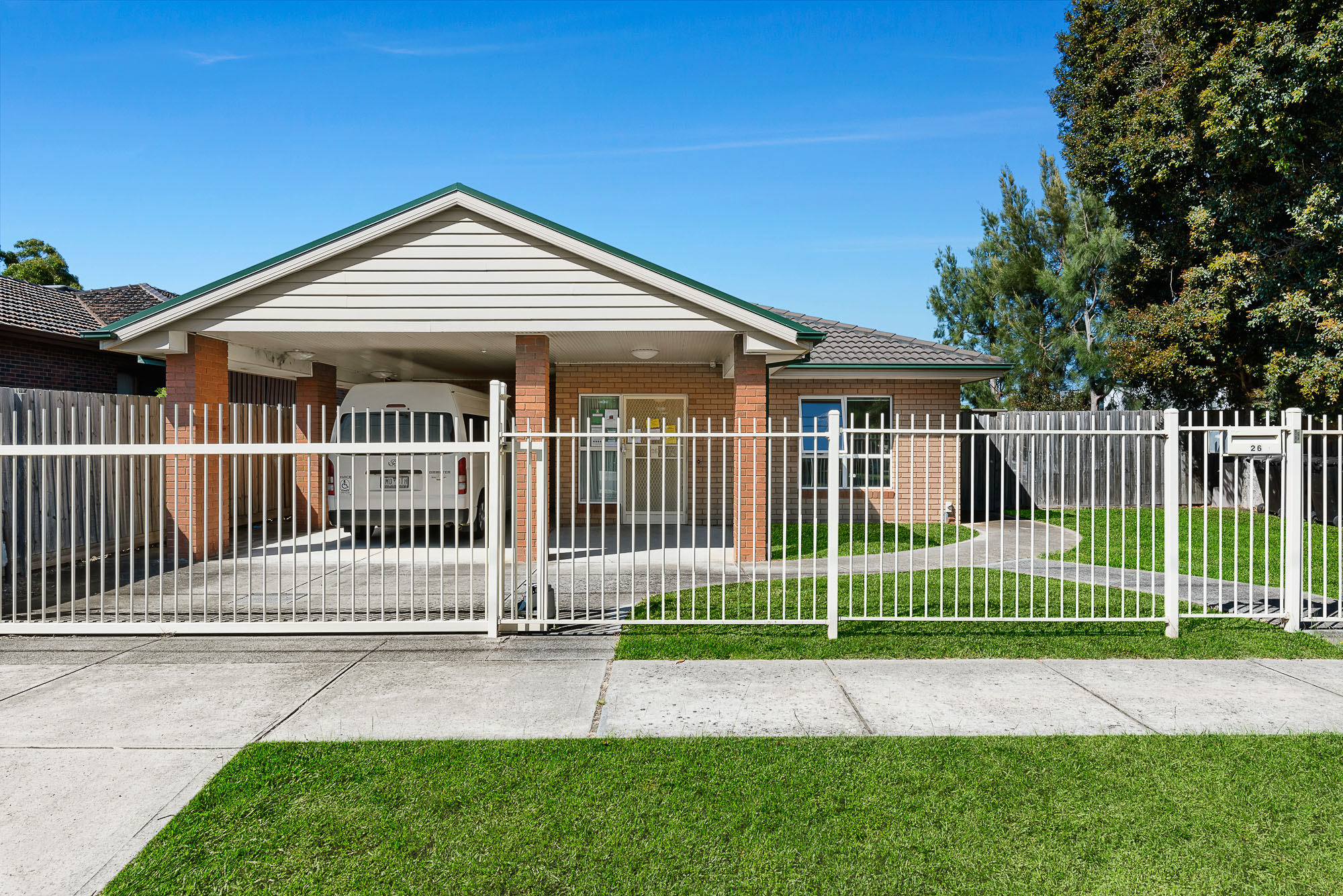 Single-storey brick home with covered carport, white fence, and paved driveway.