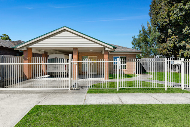 Single-storey brick home with covered carport, white fence, and paved driveway.
