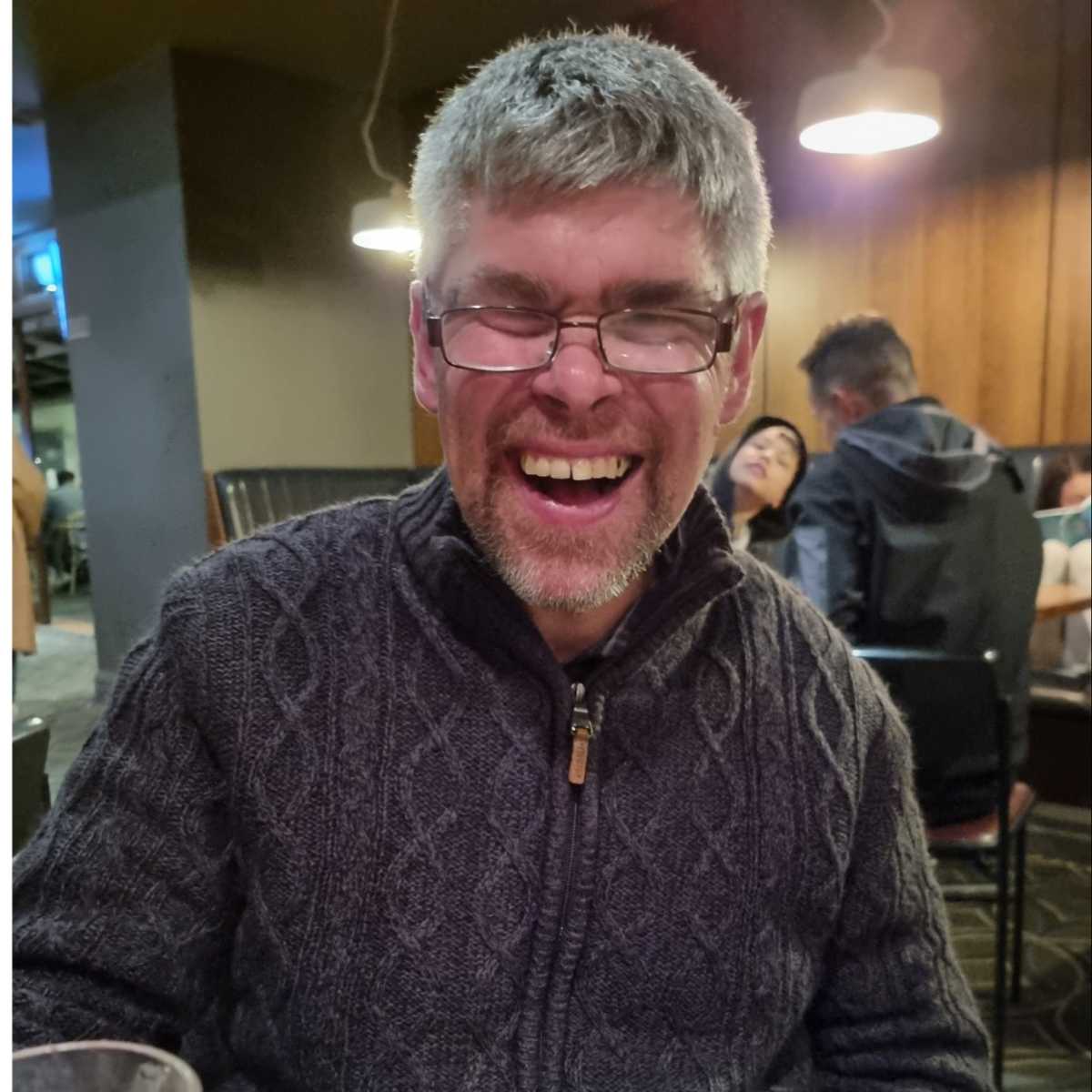 David wearing a knitted jumper and glasses is smiling at the camera.