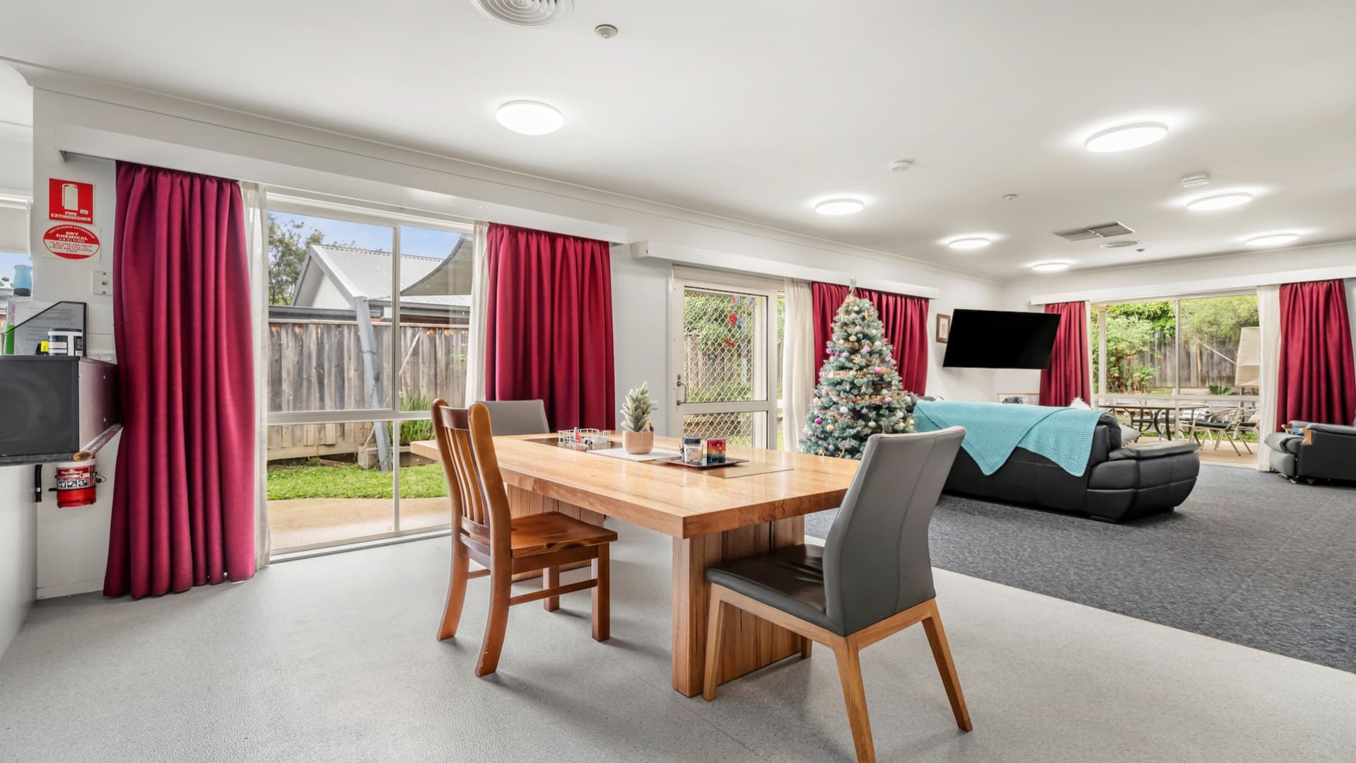 --Dining area with a large wooden table and three dining chairs. To the left is the kitchen, and to the right is the lounge room. Red curtains hang from the windows.--