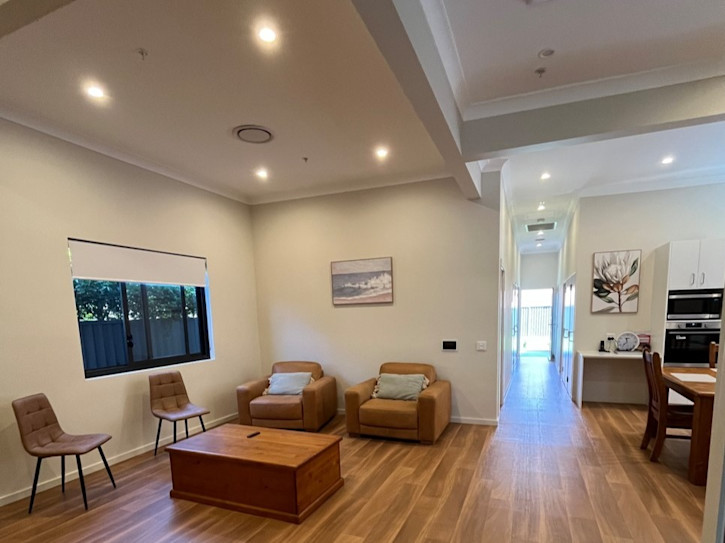 Open-plan interior view showing a lounge area with tan armchairs and a wooden coffee table, transitioning into a dining space and kitchen with wooden floors and neutral-toned walls.