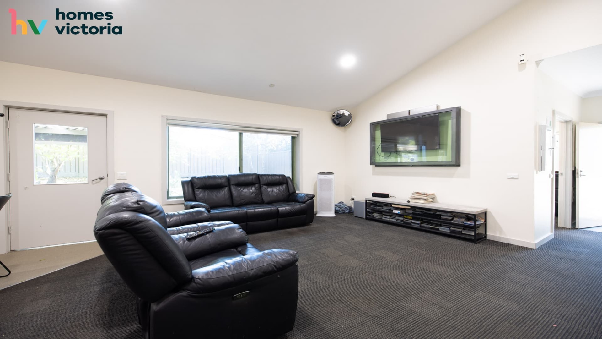 spacious lounge room with grey carpet, black leather armchairs and lounge setting facing a wall mounted television.