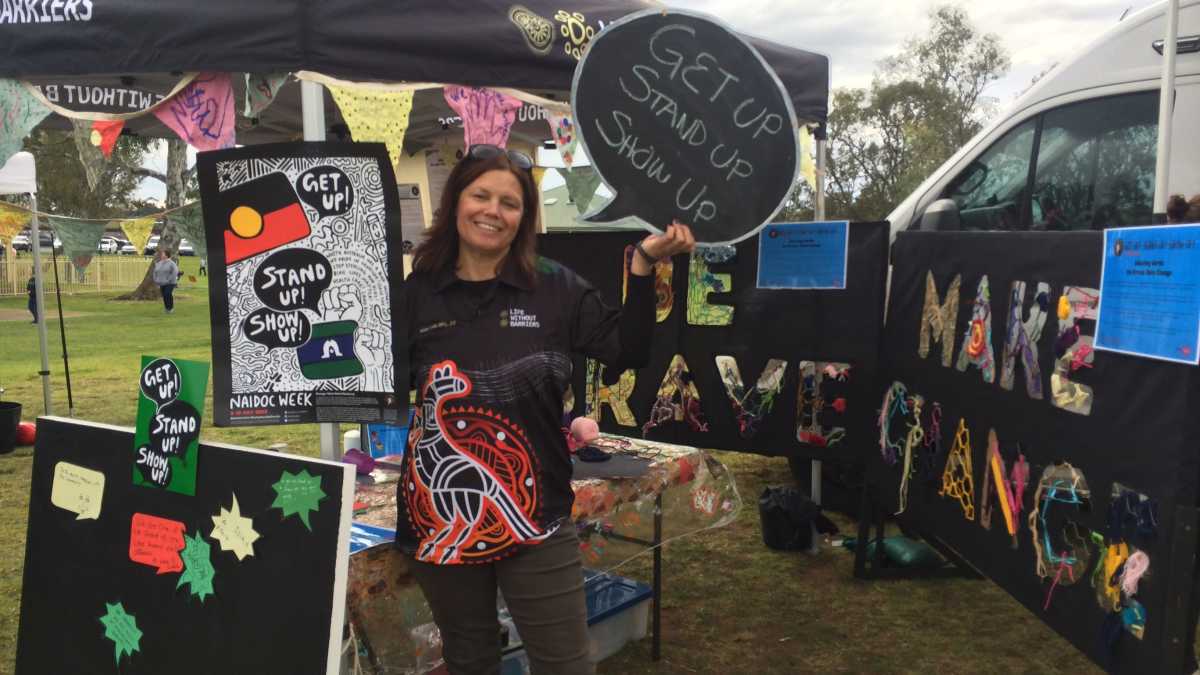 A woman swearing a Life Without Barriers black polo with an Aboriginal design on it stands in front of a stall with signs. The signs say 'Get Up, Stand Up, Show Up'