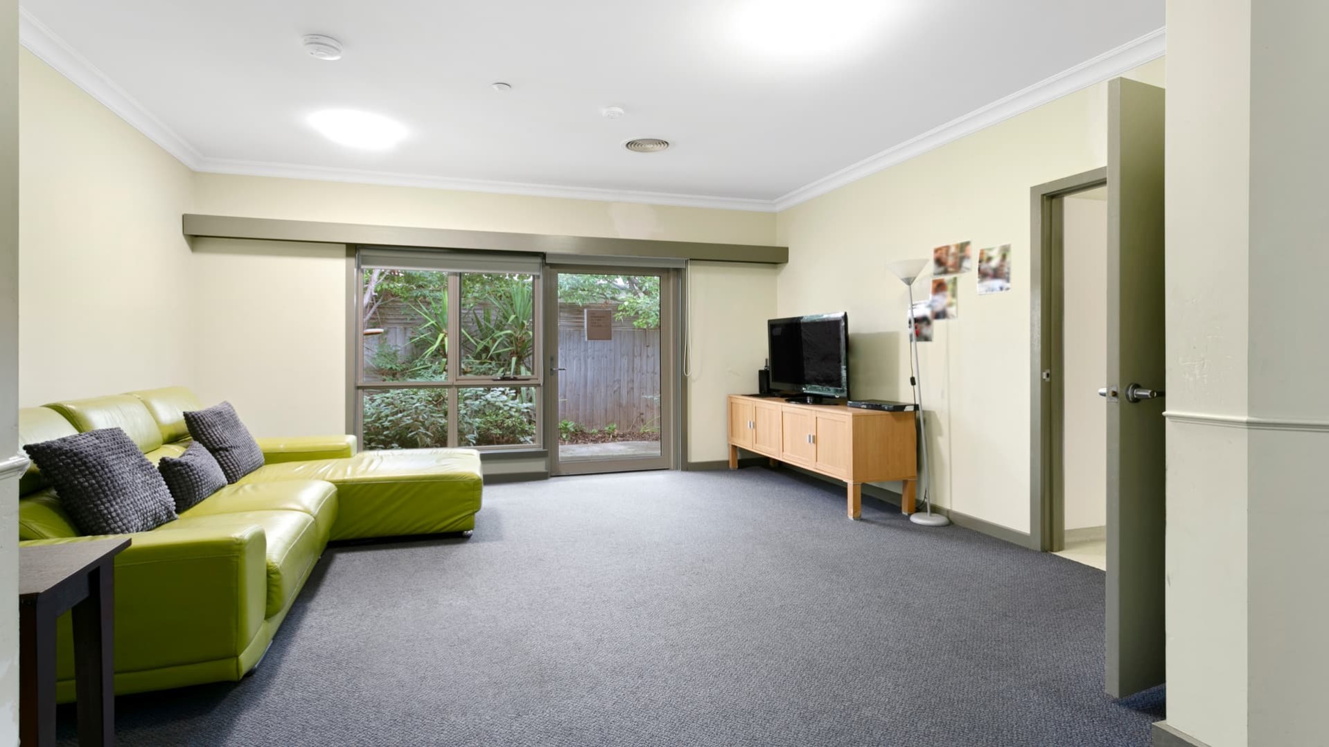 Second living space with grey carpet, three seater green lounge facing a television on a wooden cabinet.  There is a sliding door leading to outdoors.