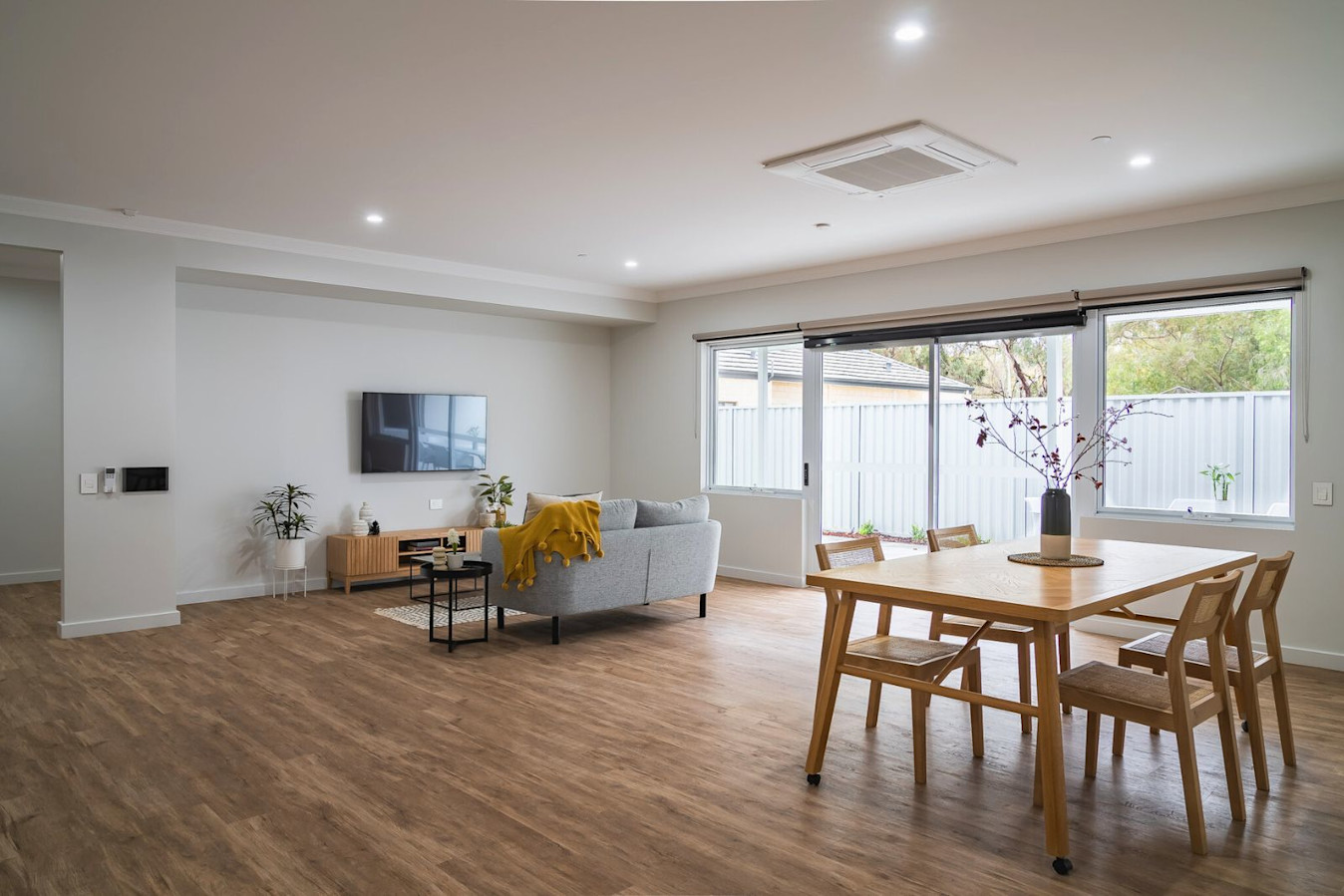 Spacious open-plan living area with wooden flooring, grey sofa, wall-mounted TV, and wooden dining table beside large windows.