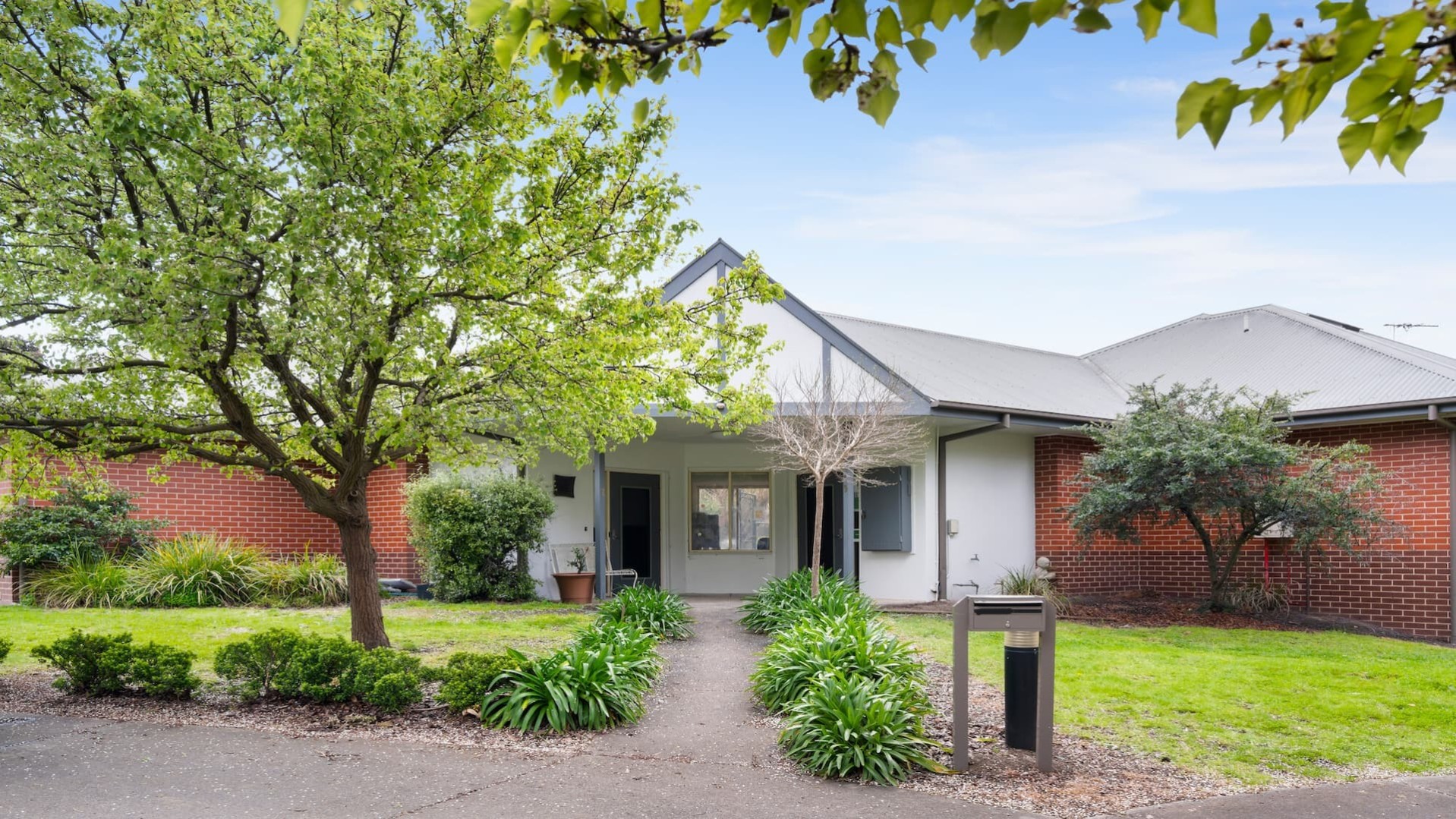 --Front of house with a tree and a concrete pathway leading to the front door. On either side of the pathway are shrubs and green grassed areas.--