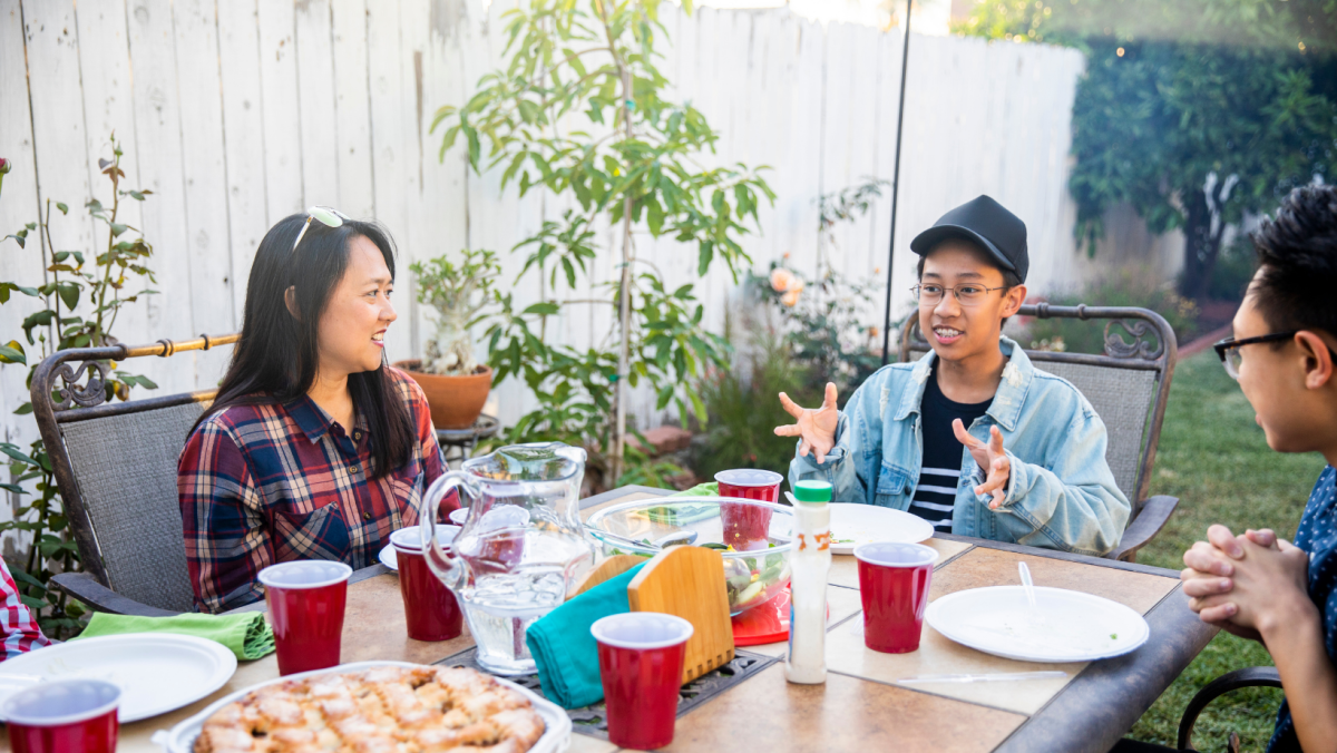 Three people sitting around a table in a backyard.. Two younger teenagers of Asian decent. One mother figure.