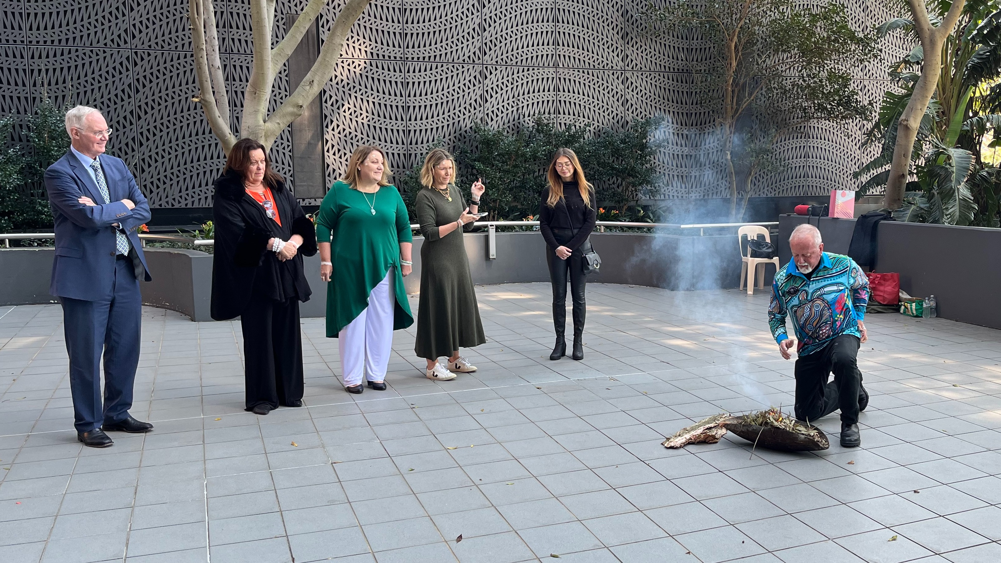 A number of people stand outdoors watching a man perform a smoking ceremony.