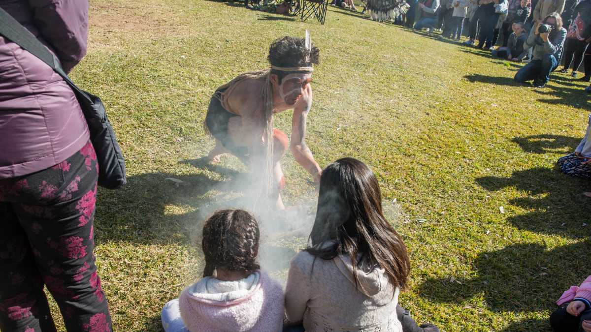 An Aboriginal man wearing face paint and a headdress performs a smoking ceremony. Two young girls sit in front of him.