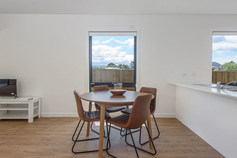 Open-plan dining area with a round table and four brown chairs, timber flooring, and large windows offering views of the outdoors and distant hills.