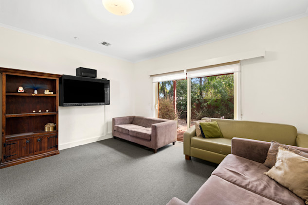 Bright living room with a large window and sliding door leading to a garden. The room features a wall-mounted flat-screen TV, a wooden shelving unit, and three sofas in muted tones of green and mauve. The space has light-colored walls, grey carpet, and minimal decor.