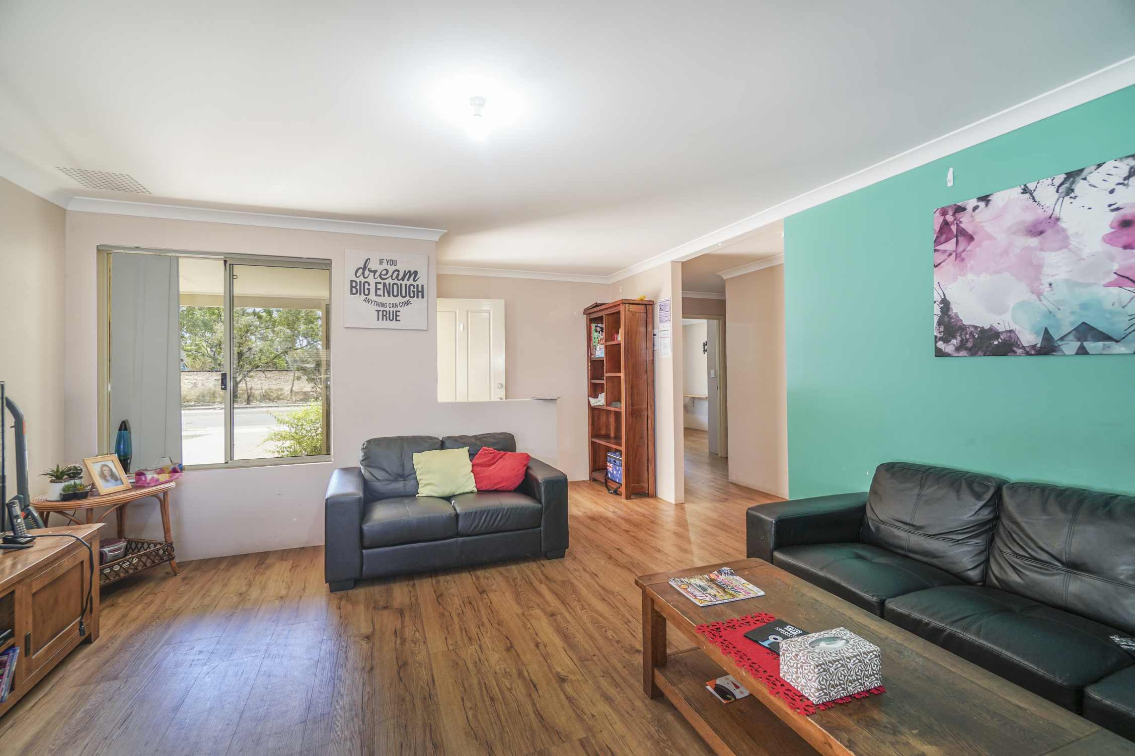 Open living room with wooden flooring, teal feature wall, dark grey couch, modern coffee table with green centerpiece, and large window overlooking the street.