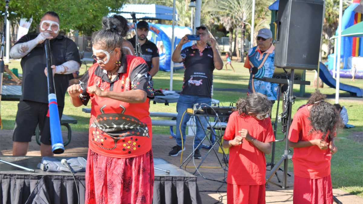 An Aboriginal woman and two children are wearing all read and dancing. Behind them is a stage with an Aboriginal man wearing white face and body paint playing the digeridoo.