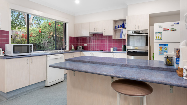 Light timber kitchen with red splashback tiles, large window overlooking garden, and blue benchtops with microwave and dishwasher.