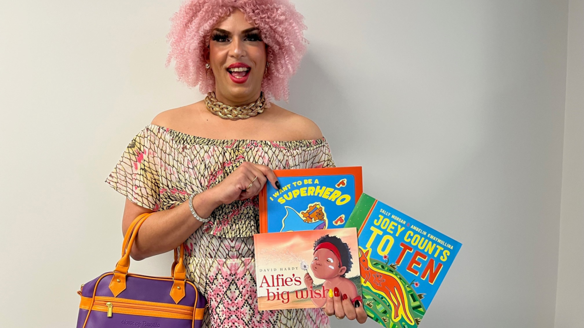 Photo of Drag Queen Estele with pink hair, a patterned dress and a purple bag holding three books.