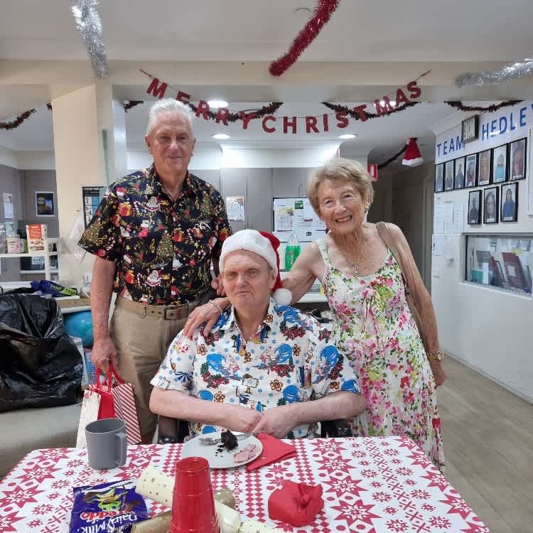 Bruce wearing a santa hat and sitting at a table decorated with Christmas decorations as two people stand behind him.