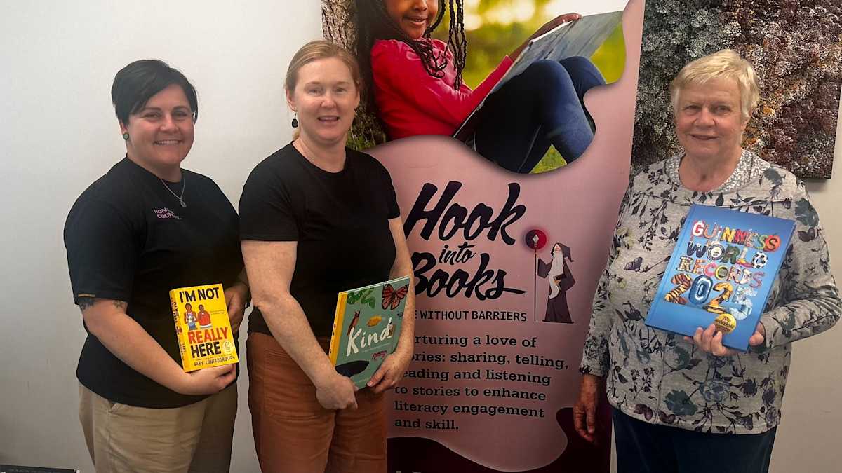 Three individuals smiling at the camera with books in their hands. Behind them is the Hook into Books™ banner.