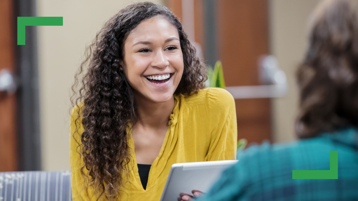 A woman with long curly brown hair wearing a yellow shirt is holding an IPad and smiling.