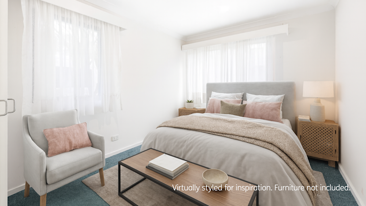 Modern styled bedroom with soft natural light, featuring a grey upholstered bed, blush and beige bedding, wooden bedside tables with lamps, an armchair with a pink cushion, and a wooden coffee table on a blue carpet.